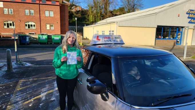 A smiling woman in a green jacket holds a flyer next to a taxi.