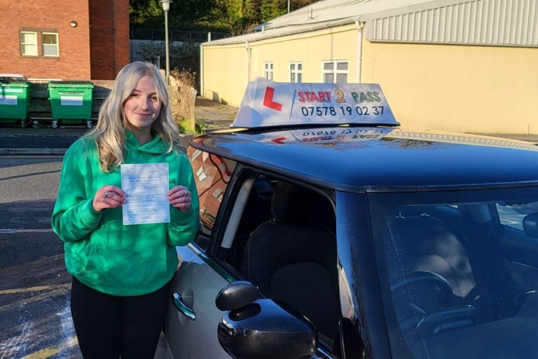 Woman smiling and holding a driving test certificate beside a learner car.