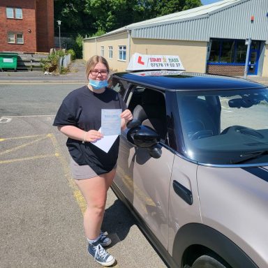 A person wearing a mask holds papers next to a driving school vehicle.