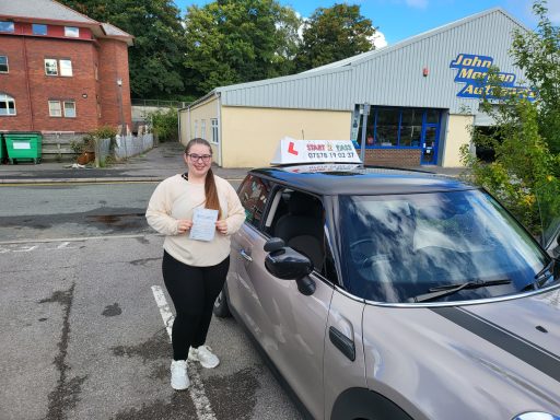 A woman stands next to a driving school car, holding a document outside a building.