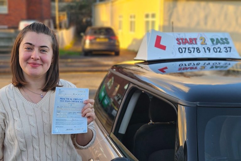 A young woman holds a driving test certificate next to a car with a learner sign.