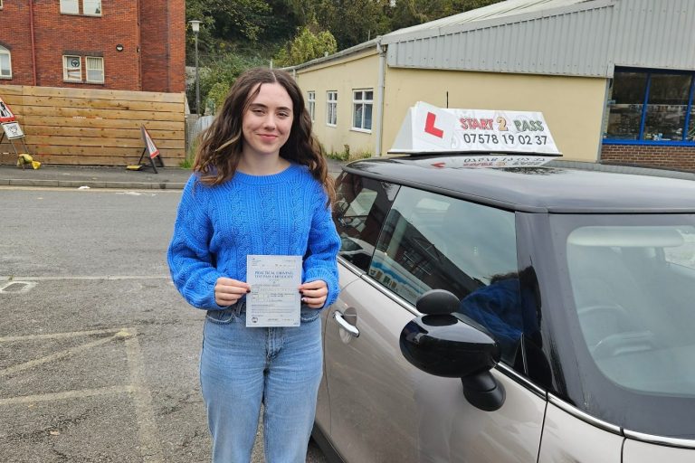 Young woman holding a driving test certificate beside a driving instructor's car.