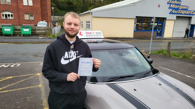 Smiling man in a black hoodie holds a certificate next to a car with a learner driver sign.