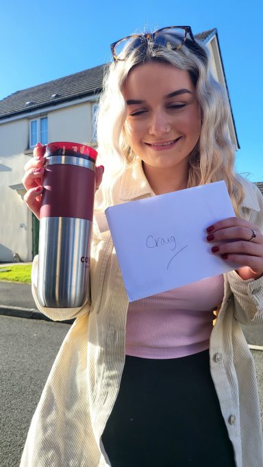 A woman smiles, holding a travel mug and a note outdoors on a sunny day.