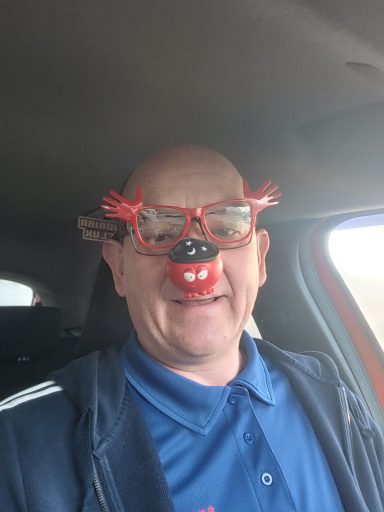 Man smiling inside a car, wearing novelty glasses and a red clown nose.