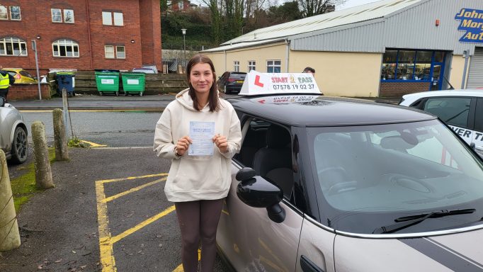 A young woman holds a certificate next to a parked driving instructor car.