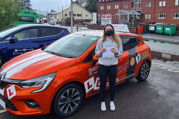 A young woman stands beside an orange driving school car, holding a certificate.