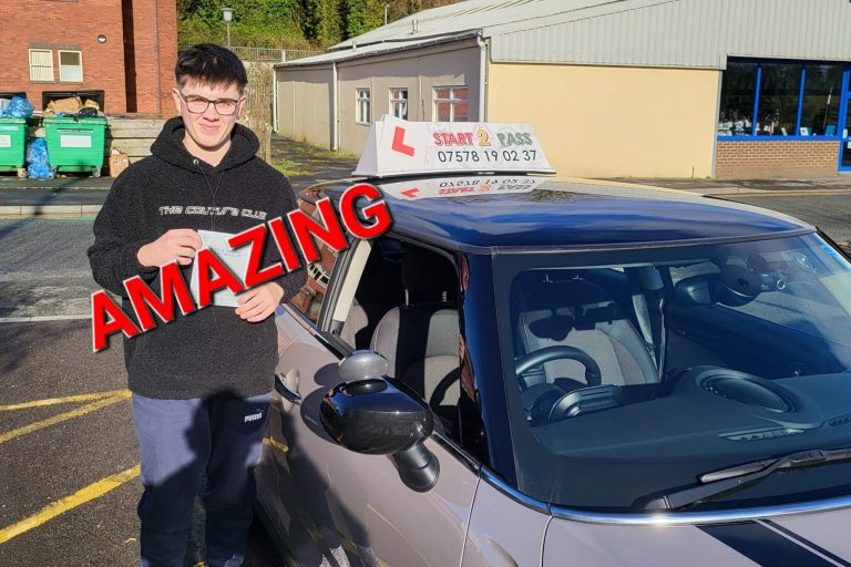 A young man stands next to a driving school car, smiling and holding a certificate.