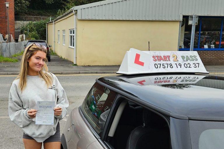 A smiling learner driver holds a test certificate next to a car with a learner sign.