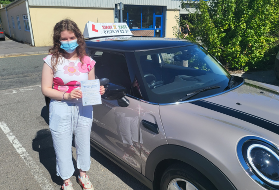 A young woman wearing a mask holds a driving test certificate next to a car.