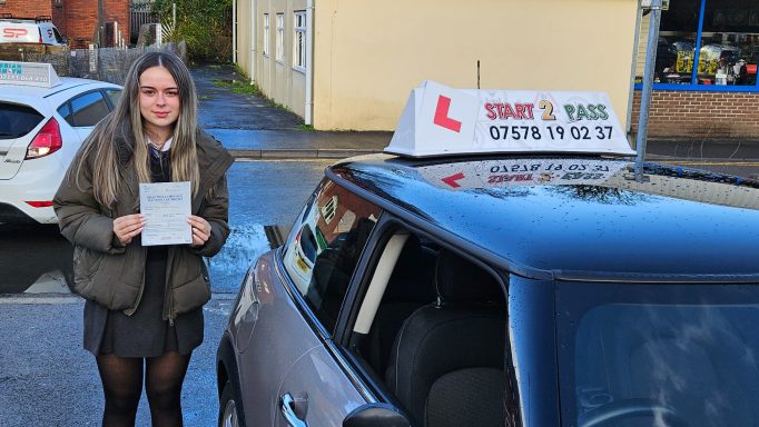 A learner driver stands next to a car with a driving school sign, holding a test pass certificate.