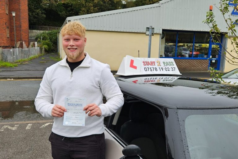 Smiling man holding a driving test certificate beside a car with a learner driver sign.