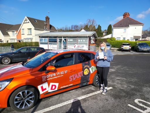 A woman stands next to an orange car with learner driver signage in a parking area.