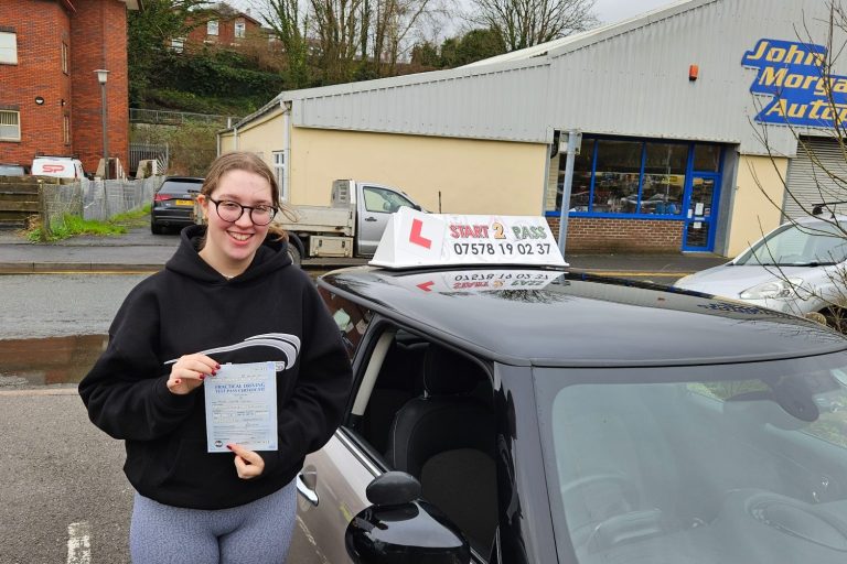 Smiling learner driver holding a certificate beside a driving school car.