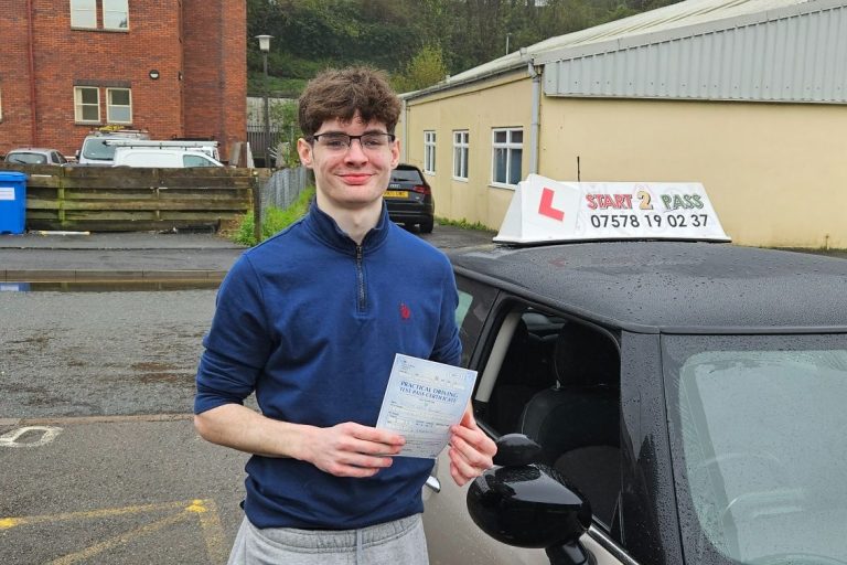 Young man smiling, holding a driving test certificate beside a driving school car.