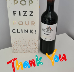 Bottle of red wine beside a decorative sign that says "Pop Fizz Pour Clink! Thank You".