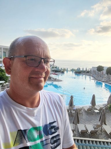 Smiling man in a white t-shirt by a pool with sea views and sunlit clouds.