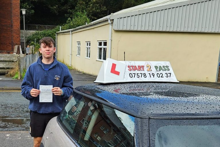 A young man stands next to a car with a driving school L-plate and holds a clipboard.