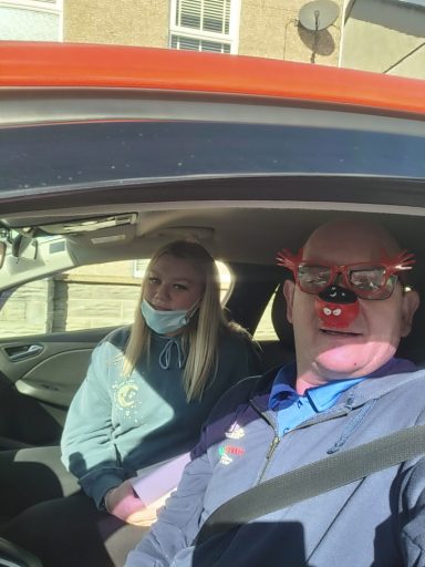 Two people in a car, one wearing a mask and the other sporting a red-nosed accessory.